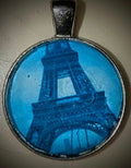 Macro photography pendant featuring a stunning blue Eiffel tower in a vintage-style decorative round frame, highlighting the delicate lace-like textures of the girders and deep blue tones.